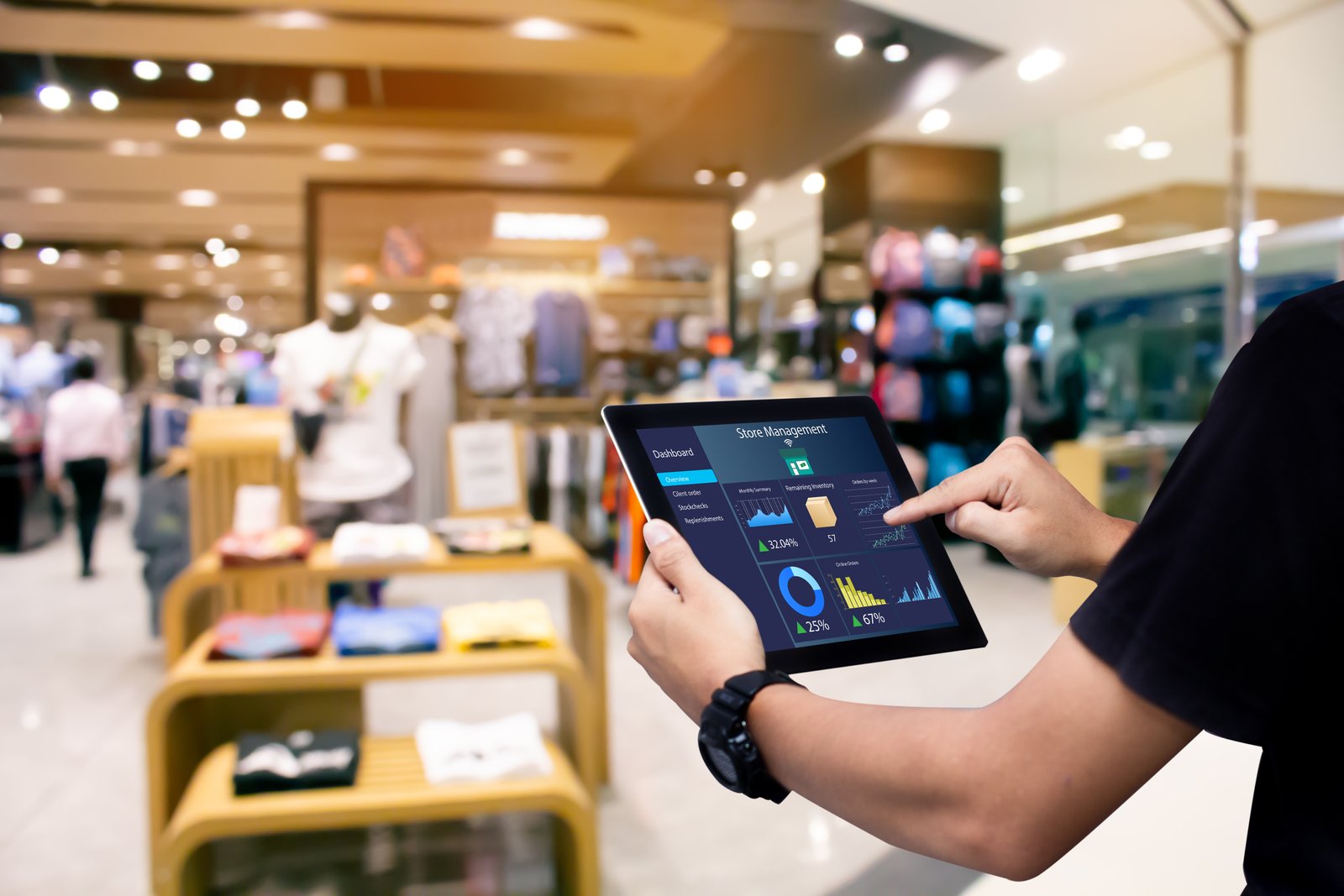 Pantalla de tablet en una tienda de ropa mostrando un tablero de control de inventario inteligente con gráficos y datos en tiempo real sobre la gestión de productos, ventas e inventarios. El fondo muestra estantes con ropa y personas dentro de la tienda, ligeramente desenfocados.