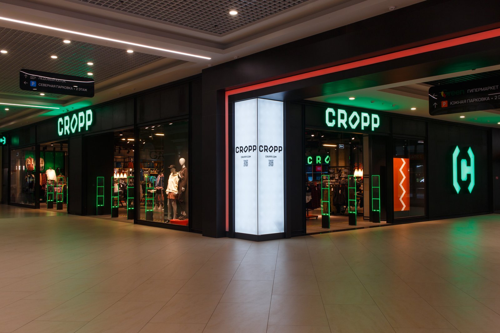 Fachada iluminada de la tienda de ropa Cropp en un centro comercial, con vitrinas que exhiben maniquíes y productos bajo una luz verde intensa. Las antenas de seguridad están integradas al diseño de la tienda, resaltando con luces verdes que se alinean con el concepto estético.